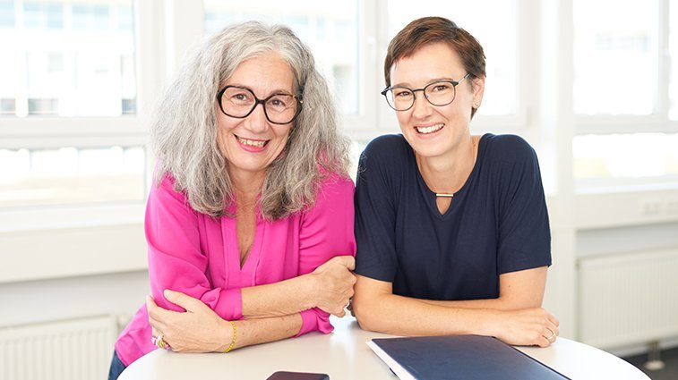 Eine Frau mit lockigen grauen Haaren steht mit einer jüngeren Frau mit kurzen brauen Haaren locker aufgestützt an einem Stehtisch.