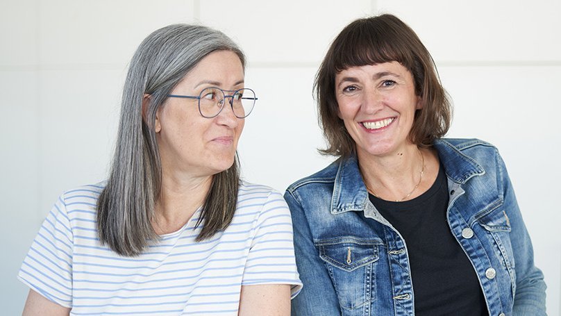 Zwei Frauen lehnen an einem Stehtisch, eine lächelt die andere an, eine lächelt in die Kamera