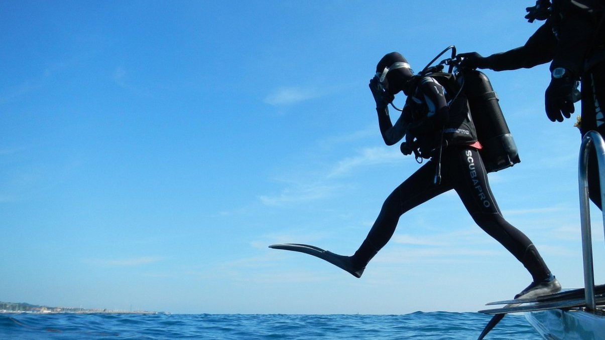 Ein Taucher in Taucheranzug mit Sauerstoff-Flasche springt ins Meer.